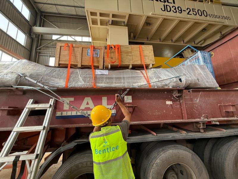 A worker wearing a yellow hard hat and a "Bentlee" high-visibility vest is tightening a metal restraint cable on a large industrial mold wrapped in protective plastic and secured with orange straps. The mold is loaded onto a red flatbed trailer inside a warehouse, with wooden cradles positioned above it for added support. An overhead crane labeled "JD39-2500" looms overhead, indicating precision handling during the loading process. This image highlights the importance of engineered securing methods tailored to the structural sensitivity of large molds.
