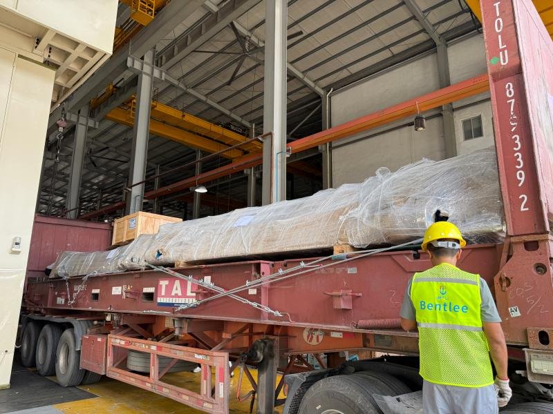 A worker in a bright yellow safety vest and hard hat stands beside a fully loaded transport trailer carrying a large mold wrapped in protective film and secured with multiple tie-down cables. The mold rests on a custom cradle within a red TAL International flatbed trailer, located inside a spacious industrial facility equipped with overhead cranes and structural steel beams. The scene emphasizes the meticulous preparation required for safe mold transportation, including proper packaging, load distribution, and alignment checks—critical steps that distinguish precision mold logistics from standard heavy equipment hauls.