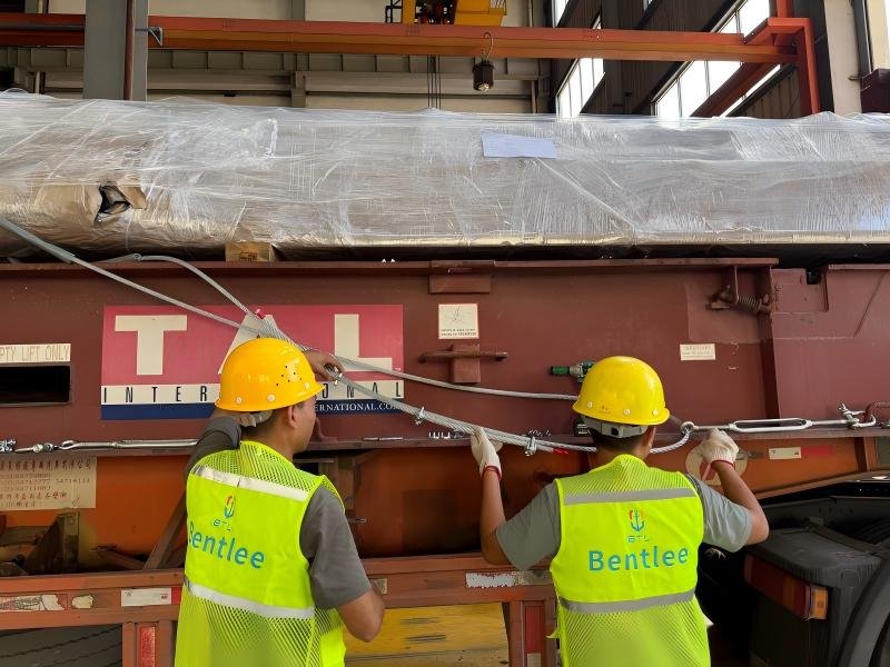 A large industrial mold covered with protective film is being loaded onto a specialized transport trailer using a XINGLI overhead crane inside a factory facility. Workers in safety gear supervise the operation, ensuring secure placement. The image illustrates critical stages of mold transportation where structural integrity and precise alignment are maintained through engineered equipment and planning.