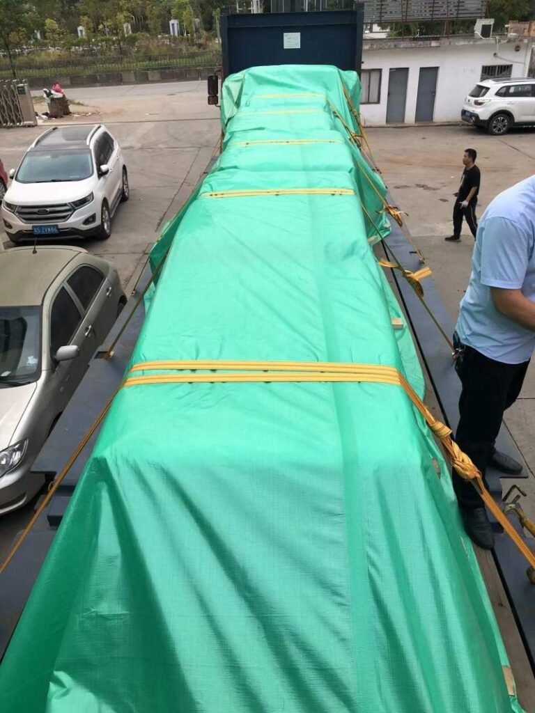 Large industrial mold covered with green tarp and secured with yellow straps on a flatbed trailer, ready for transport. Workers inspecting before departure.