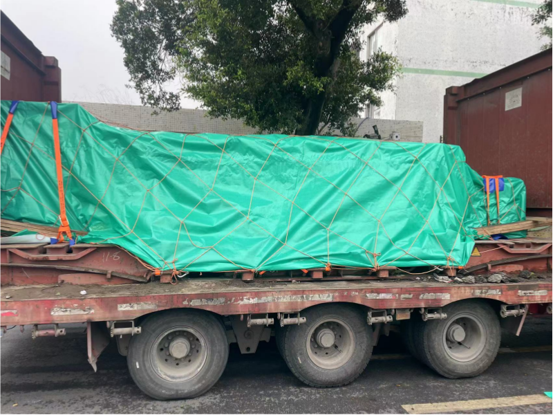 Flatbed trailer carrying a large mold covered with green tarpaulin and secured with netting, parked outdoors before shipment.