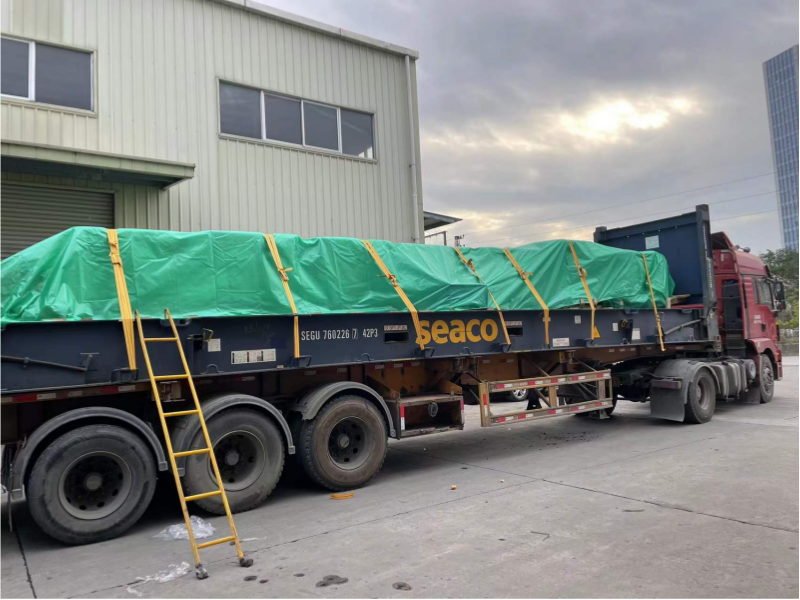 Lowboy trailer with green tarp and yellow straps parked outside a factory, labeled SEGU 760226, ready for long-haul mold shipping.