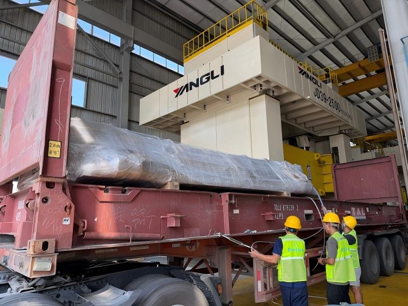 Three workers in high-visibility vests and yellow hard hats are securing a large industrial mold wrapped in protective plastic onto a red flatbed trailer inside a warehouse. One worker stands on a ladder adjusting the load, while two others monitor the process. The scene highlights proper handling procedures during mold transportation to prevent damage.