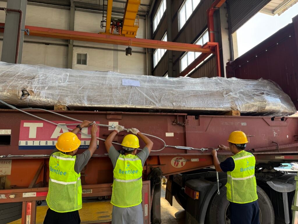 Workers installing wire rope lashings on oversized cargo loaded onto a flat rack trailer in a warehouse