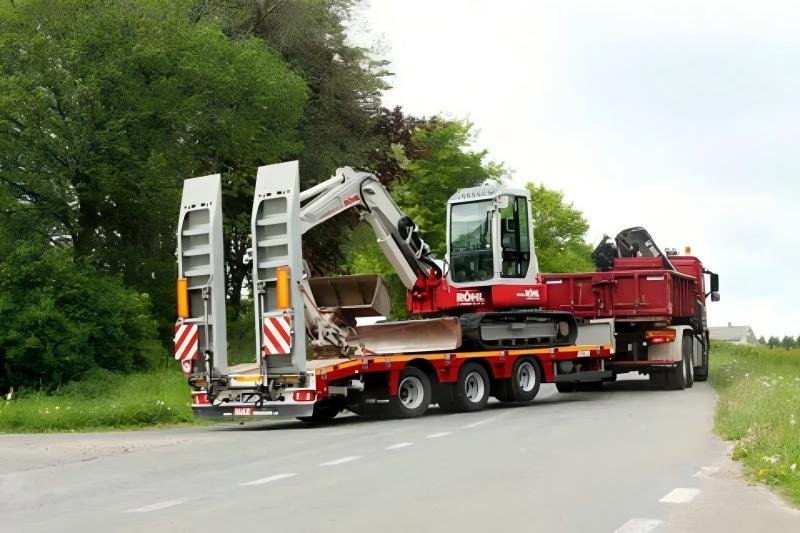 A red lowboy semi-trailer safely transporting a large hydraulic excavator on a public road, demonstrating professional engineering logistics capabilities.