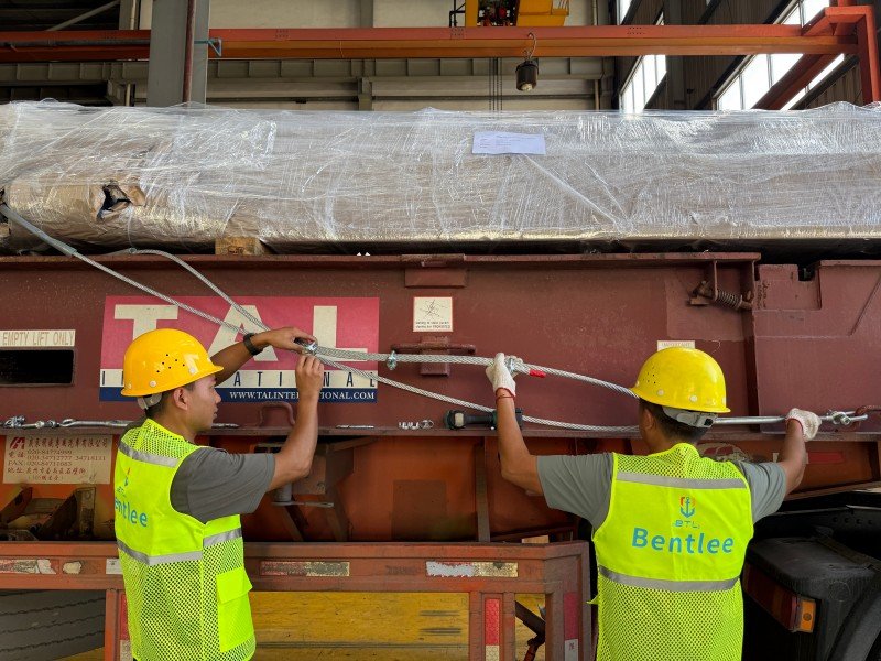 Two workers tightening wire rope lashings with turnbuckles on a flat rack to restrain heavy industrial equipment against lateral and longitudinal forces.