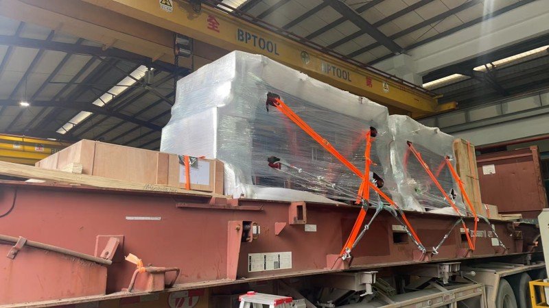 Large industrial component wrapped in plastic and secured with multiple orange lashing straps on a flat rack container