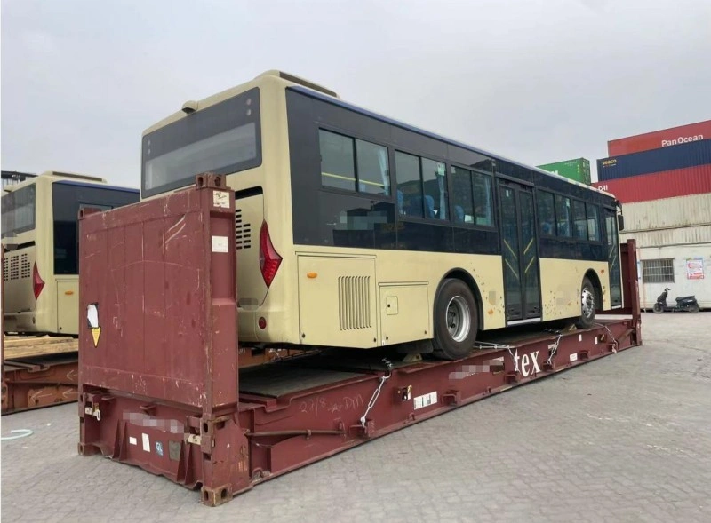A beige and black city bus is securely positioned on a red flat rack container at a port yard, with visible tie-down chains and structural supports ensuring stability during transport.