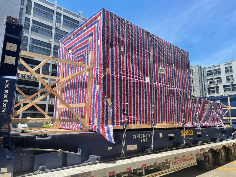 Large over-height OOG cargo secured on an open top container with red-white-blue tarpaulin, timber blocking, and chain lashing systems ready for sea transport.