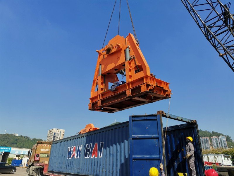 Orange heavy machinery being lifted by crane into blue open top container with workers supervising the operation