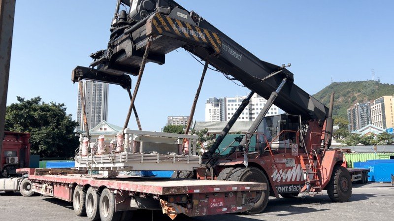 Industrial machinery being lifted by reach stacker onto flatbed trailer using certified lifting points and rigging
