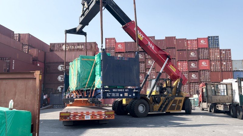 Yellow reach stacker lifting oversized cargo covered with green tarpaulin onto blue open top container at port terminal