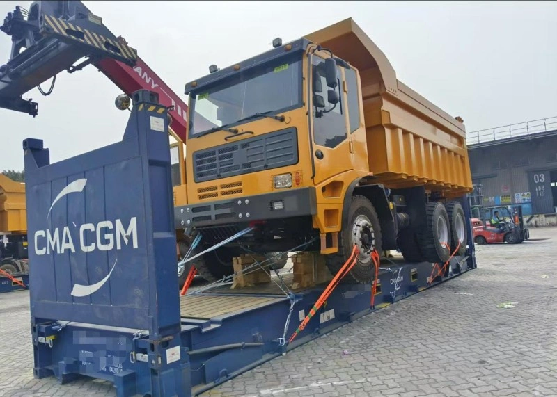 A large yellow dump truck is being loaded onto a blue CMA CGM open top container using a mobile crane, with orange lashing straps and wooden dunnage securing the vehicle for sea transport.