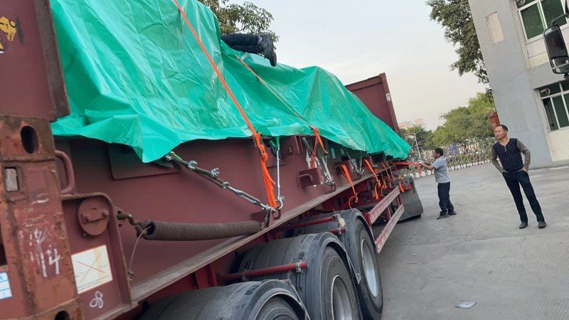 Heavy equipment secured with orange straps and metal chains on flat rack trailer during loading preparation