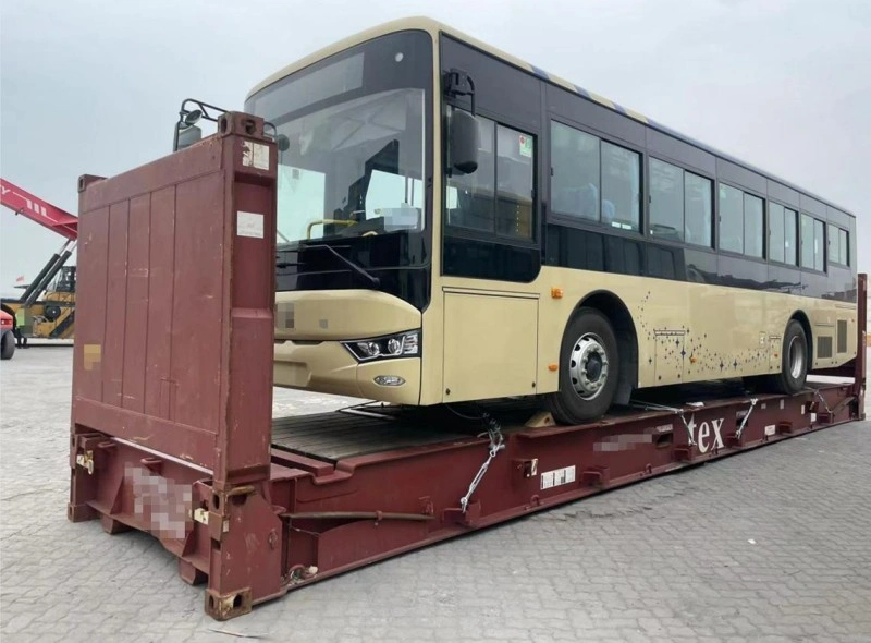 Front-side view of a beige city bus on a flat rack container, highlighting the front axle placement, cabin structure, and securing hardware used to comply with international cargo safety standards.