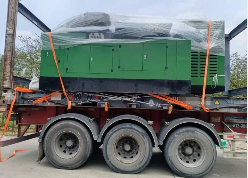 A large green generator set, partially wrapped in clear plastic film and strapped with orange tie-downs, is mounted on a multi-axle heavy-duty trailer for oversized freight transport.