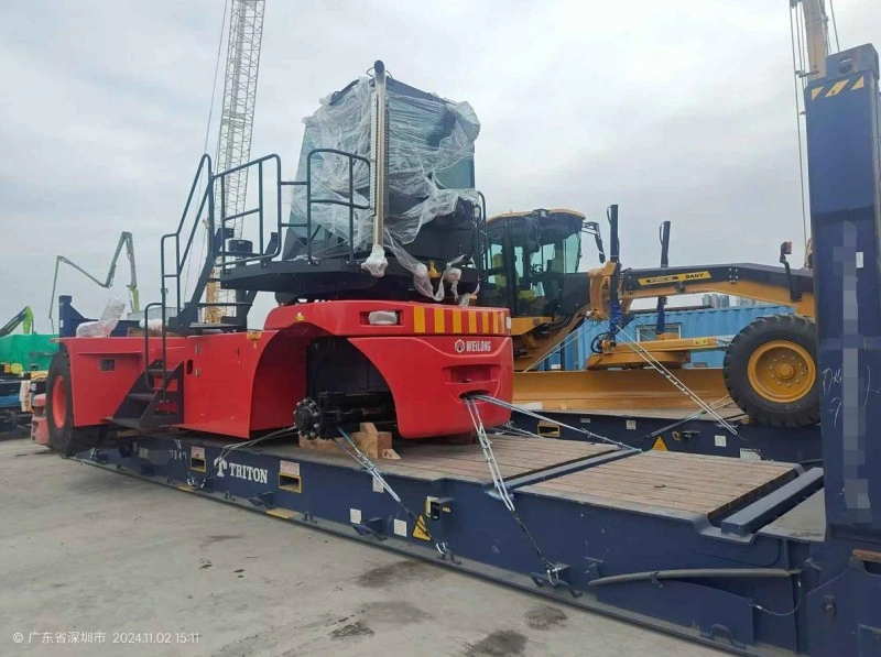 A red Weelong reach stacker and a yellow motor grader are secured with steel chains and wooden blocks on a blue Triton flat rack container at a port terminal, with cranes visible in the background.