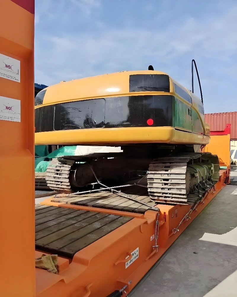 Front view of a large yellow tracked excavator properly positioned on wooden dunnage inside an open-top flat rack container, ready for lashing.