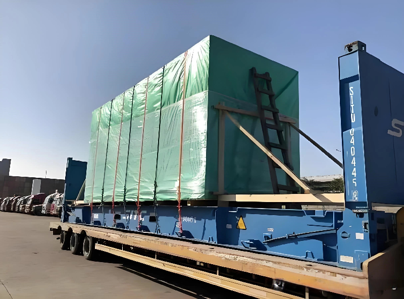 A blue flat rack container loaded with green-wrapped oversized machinery secured by multiple tension belts, parked in a logistics yard with a fleet of trucks in the background, ready for route execution.