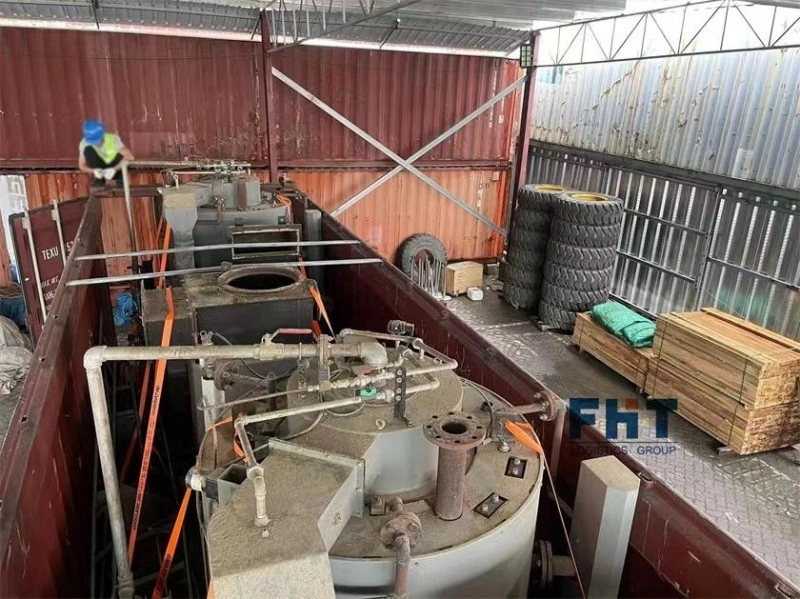 A worker in a blue hard hat and safety vest inspecting heavy industrial equipment loaded in an open top container, with wooden dunnage, tires for stabilization, and orange lashing straps.