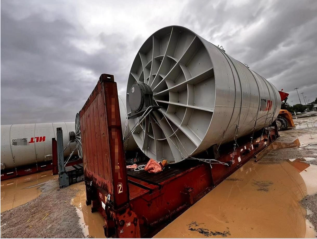 A large, cylindrical industrial fan with a prominent blade assembly is loaded onto a red flatbed trailer, secured with chains, in a muddy yard under an overcast sky.