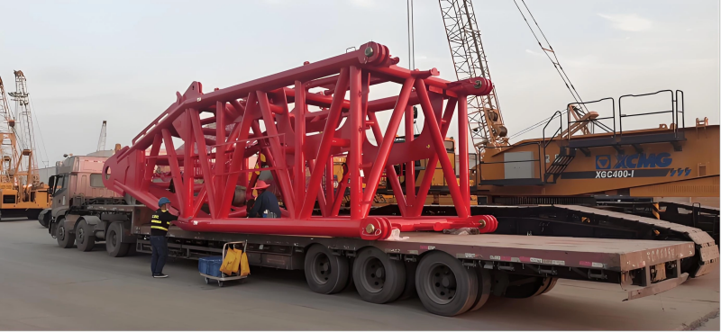 A massive red lattice boom section of a crawler crane being transported on a heavy-duty multi-axle low-bed trailer at a port facility, illustrating the logistics of moving oversized cargo that requires specialized equipment and route planning.