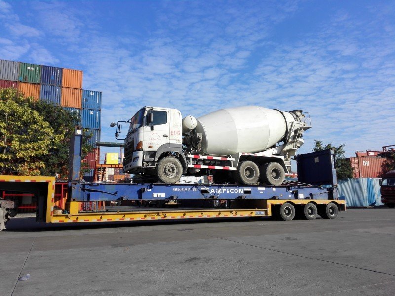 Concrete mixer truck being lifted onto lowboy trailer for OOG cargo shipment in a container yard ✅ Caption:
