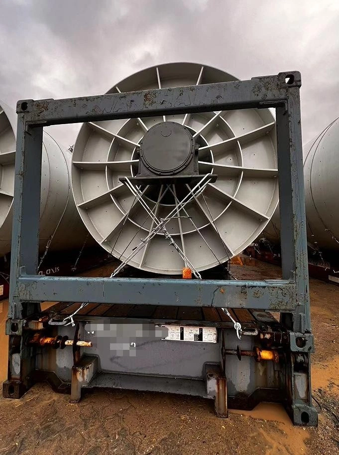 A large industrial axial fan with a grey metal casing and radial blades is securely strapped to a blue flat rack container, ready for heavy-lift shipping at an outdoor logistics yard.