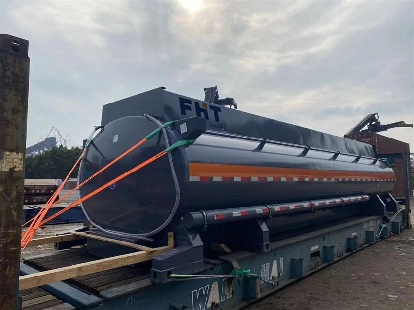 A large grey cylindrical industrial tank with an orange stripe is strapped down with orange and green tie-downs onto a blue flat rack container, ready for international transport.