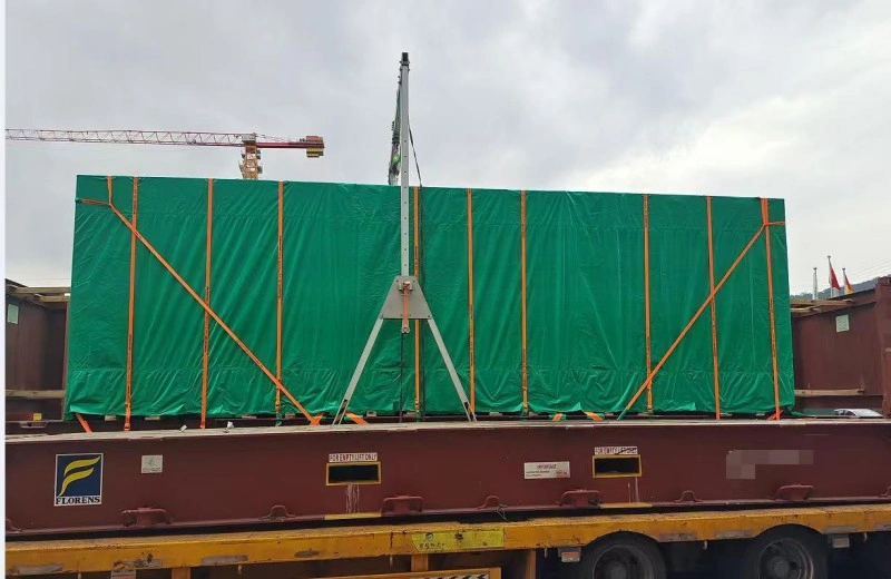 A large cargo unit wrapped in green tarpaulin and secured with orange lashing straps is being lifted by a mobile crane onto a flat rack container at a port terminal, with construction cranes visible in the background.