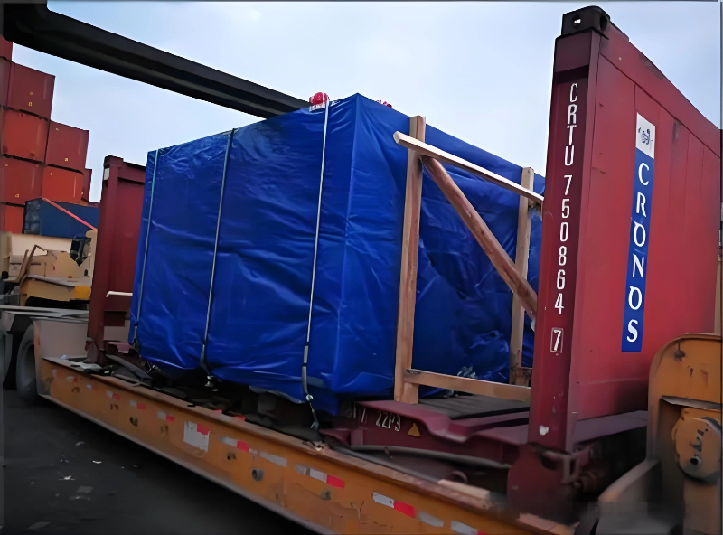 A large cargo unit covered in blue tarpaulin is secured on a red flat rack container using wooden beams and steel straps at a shipping terminal, highlighting the preparation phase of international route planning.