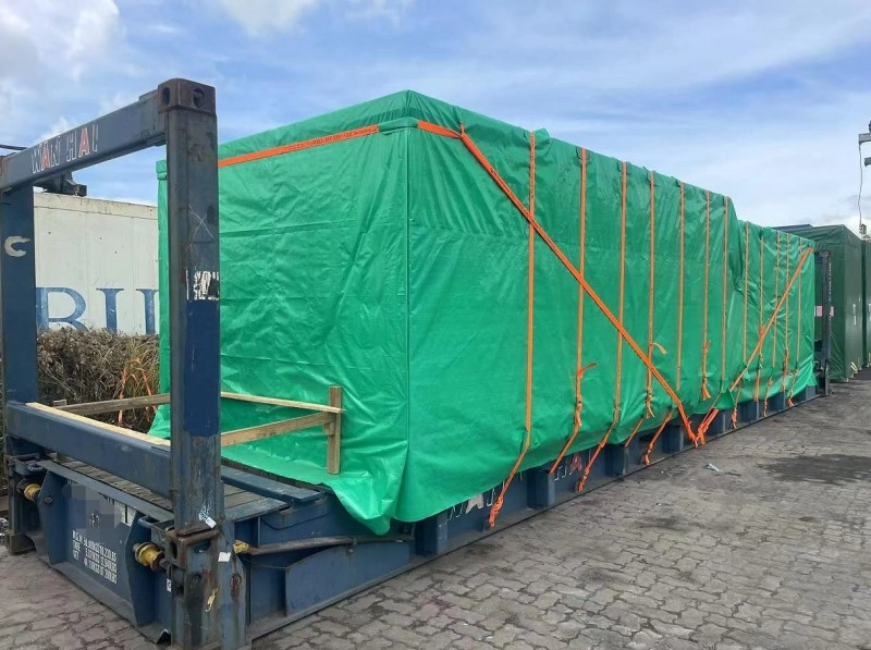 An elongated piece of machinery covered in green tarpaulin and secured with multiple orange straps is loaded onto a blue flat rack container at a port yard.
