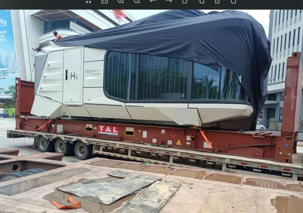 A white modular cabin unit with large glass windows is partially covered by a black tarp and secured on a red flat rack container, with visible orange tie-downs and structural supports ensuring stability during transport.