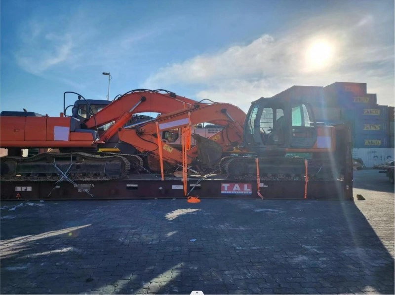 Two orange excavators are loaded closely together on adjacent flat rack containers in a sunny port yard, with shipping containers stacked in the background.