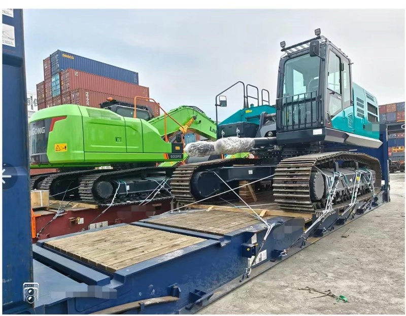 Two excavators, one green and one teal, are positioned side-by-side on separate flat rack containers, with shipping containers stacked in the background at a busy port.