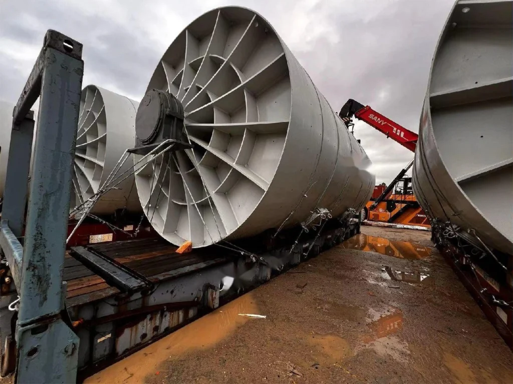 Several large, grey industrial fans are staged on flat rack containers in a muddy yard, with a red SANY heavy-lift crane positioned for loading operations.