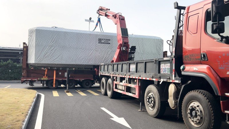 Red truck with crane arm unloading large cargo covered in gray tarpaulin from flatbed trailer at logistics yard