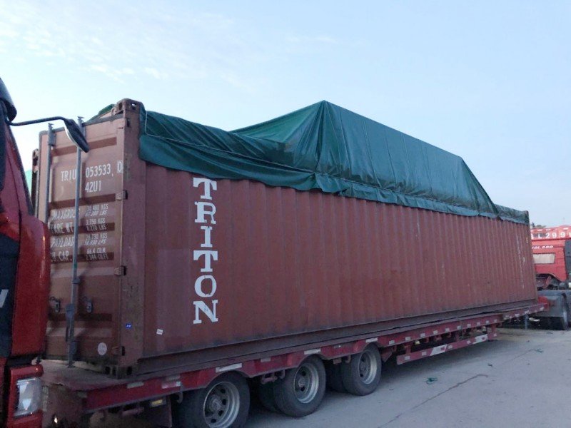 Triton-branded open top container covered with green tarpaulin on flatbed trailer during transport