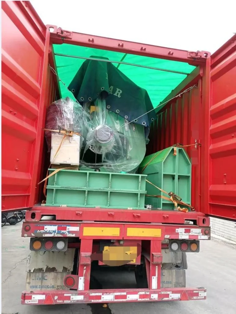 A red open top shipping container with its doors open, revealing a large green industrial machine wrapped in plastic and secured with orange lashing straps inside, loaded onto a red flatbed trailer.