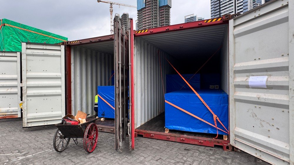 Open top container being loaded with a tall piece of heavy equipment, covered with a blue tarp, at a shipping yard with cranes in the background