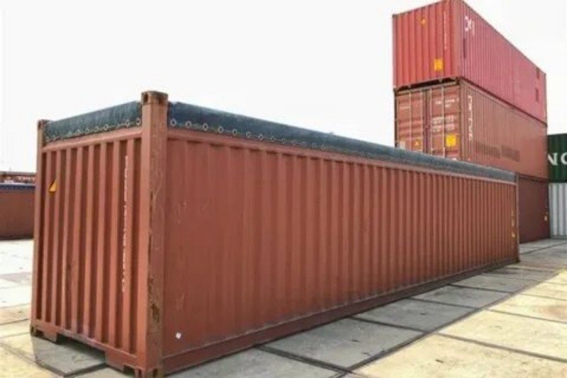Red open top shipping container with tarpaulin cover sitting in a terminal yard near stacked dry containers, illustrating port special handling charges and trade lane equipment availability issues