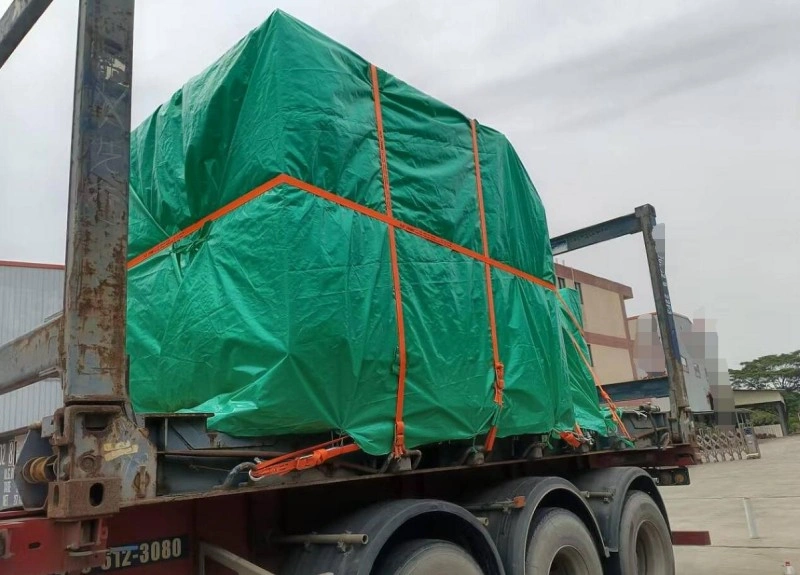 A large industrial machine wrapped in green waterproof tarpaulin is strapped with orange tie-downs onto a flatbed trailer, ready for oversized cargo shipping.