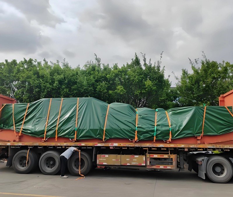 Large industrial equipment covered with green tarpaulin and strapped to a flatbed truck, demonstrating breakbulk shipping methods for oversized cargo that cannot fit in standard containers.