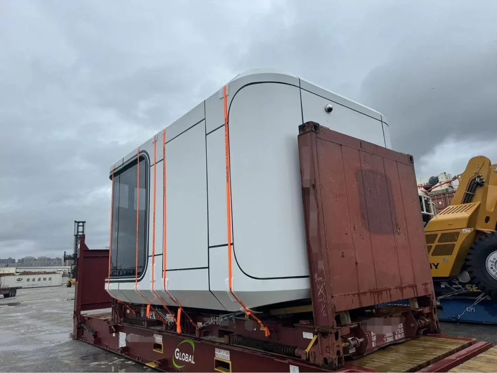 A white rectangular prefab module with rounded corners and large side windows is strapped with orange tie-downs onto a brown flat rack container at a port yard under overcast skies.