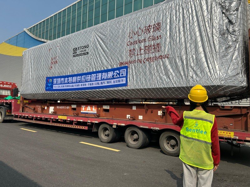 Logistics worker in yellow safety vest inspecting large cargo covered with silver tarpaulin on flatbed trailer at industrial site