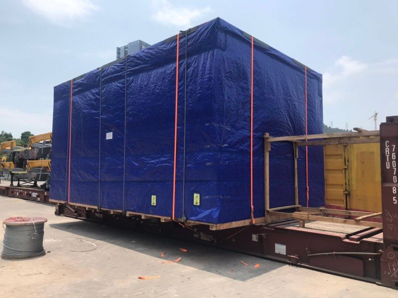 Large industrial equipment covered with blue tarpaulin and secured with orange straps on a flat rack trailer