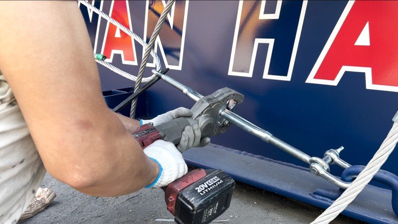Worker using electric hydraulic wrench to tighten steel cable fittings on mining machinery during container loading
