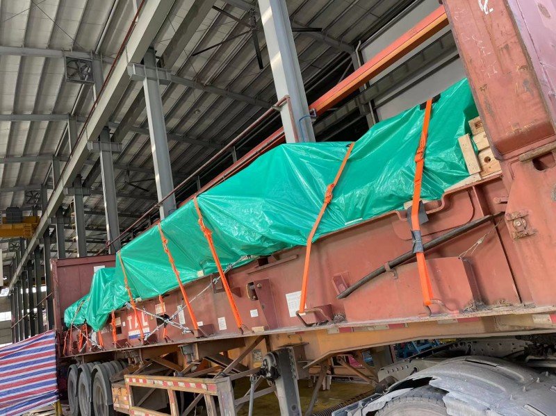 Construction equipment covered with green tarpaulin and secured with orange straps on flatbed trailer inside warehouse
