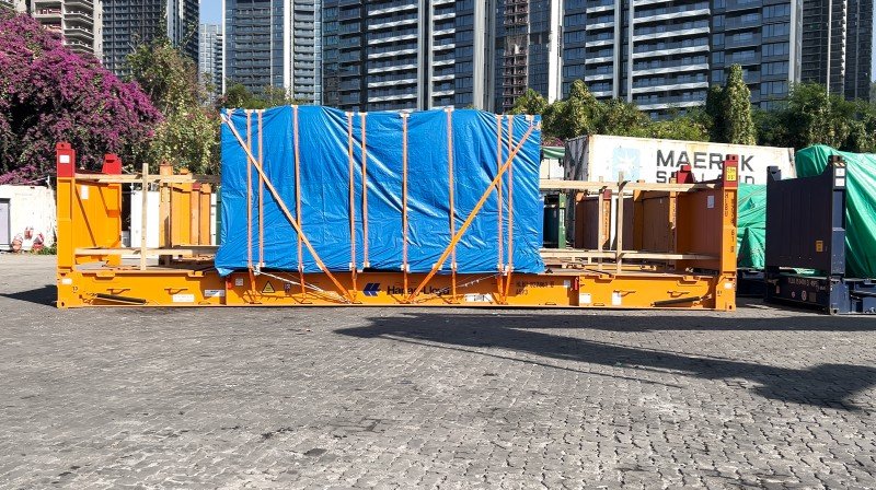 Large industrial machinery covered with blue tarpaulin and secured with orange straps inside open top container for safe transportation