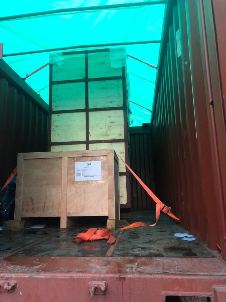 Wooden crate containing hazardous materials secured with orange straps inside open top container under green tarpaulin cover